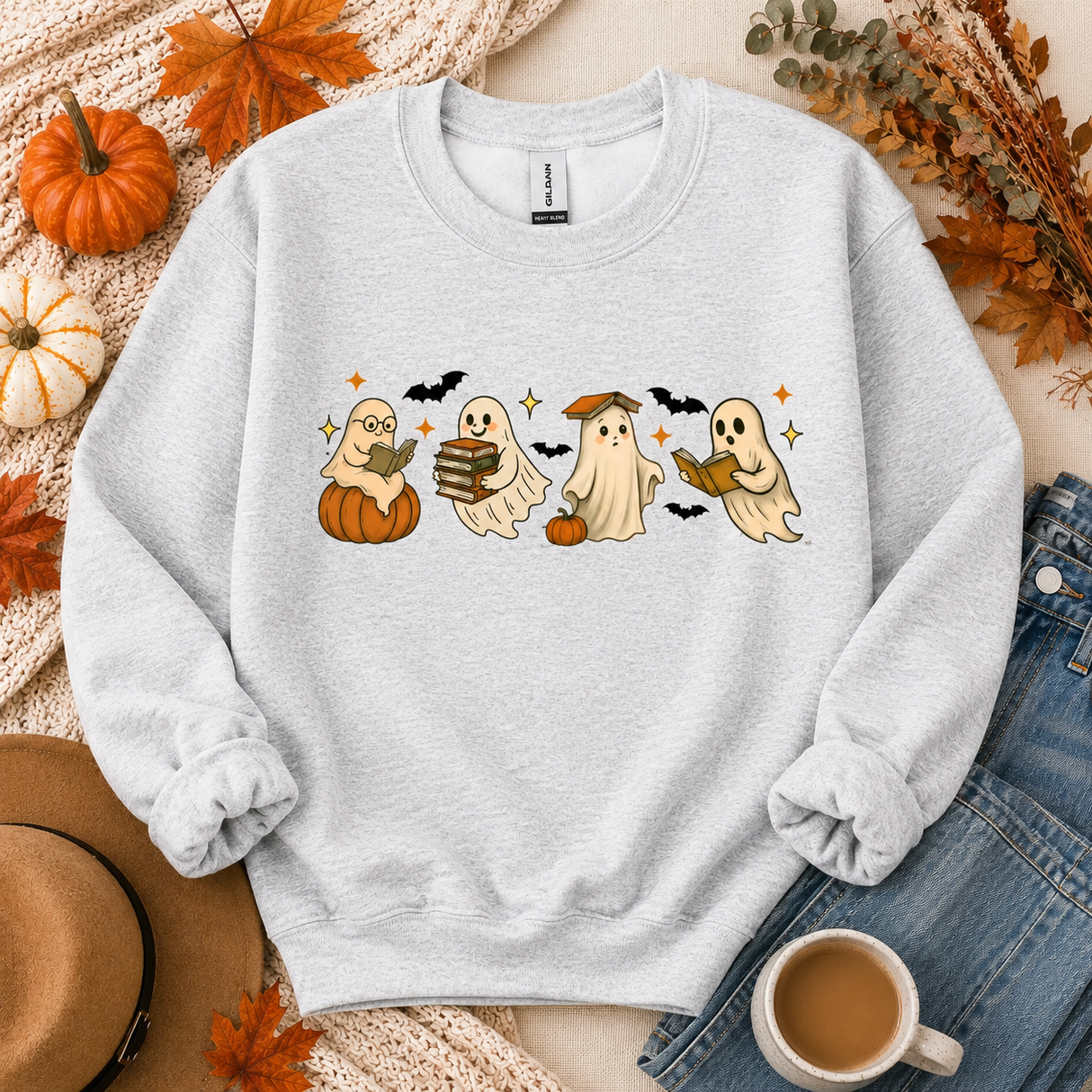 A light heather gray crewneck sweatshirt featuring a cute cartoon graphic of four ghosts reading books with bats and pumpkins. The sweatshirt is styled in a flat-lay arrangement with autumn props including pumpkins, a hat, a cup of coffee, and denim jeans.