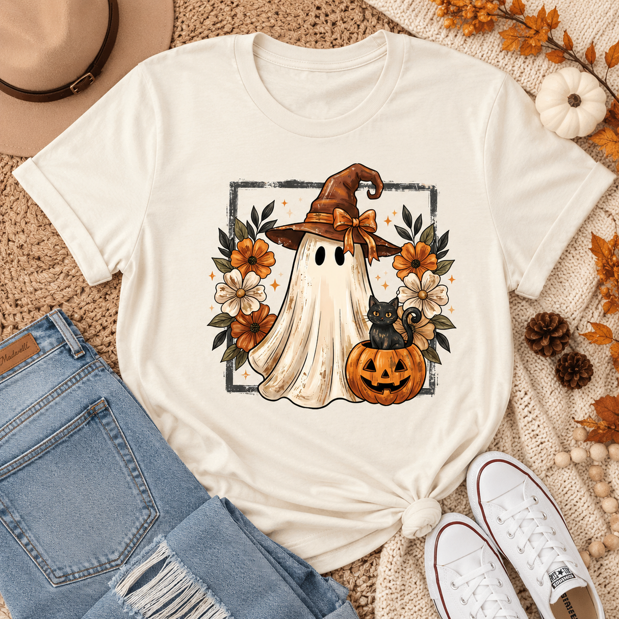 An off-white graphic t-shirt featuring a cute Halloween design of a ghost in a witch hat with a black cat and jack-o'-lantern, styled in a flat lay with jeans, white sneakers, a hat, and autumn leaves on a beige knit blanket.