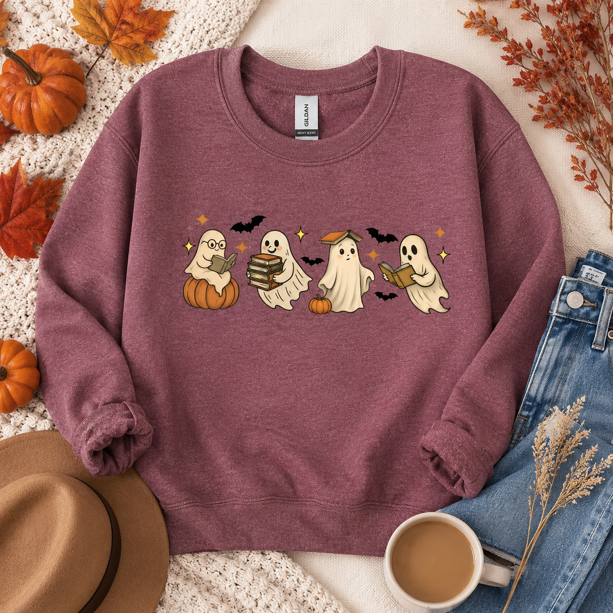 A mauve crewneck sweatshirt with a graphic of four cute cartoon ghosts reading books, styled in a flat lay with autumn leaves, pumpkins, a hat, jeans, and a cup of coffee.