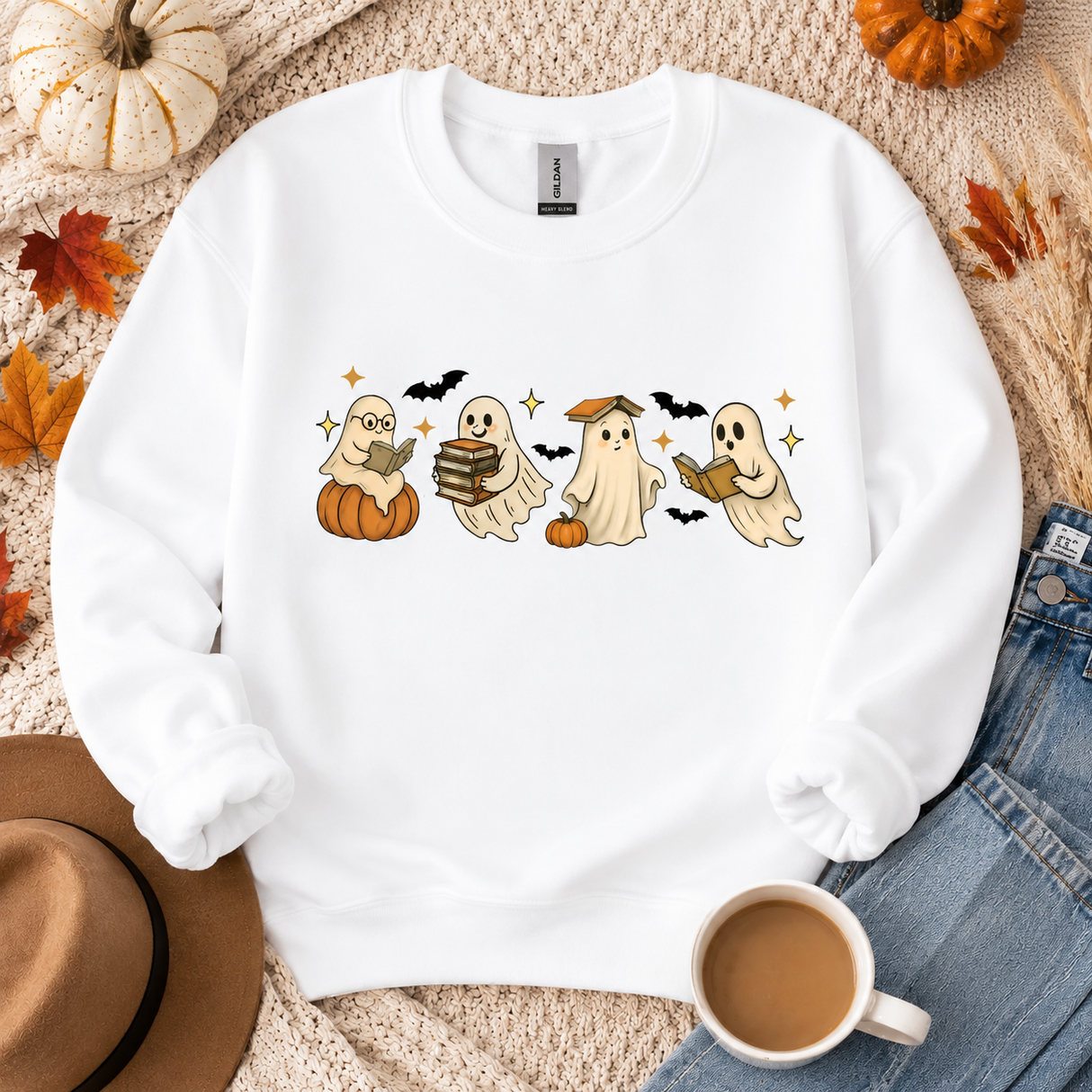 A white crewneck sweatshirt with a cute Halloween graphic of four ghosts reading, styled in a flat lay with autumn leaves, pumpkins, a hat, jeans, and a coffee mug.