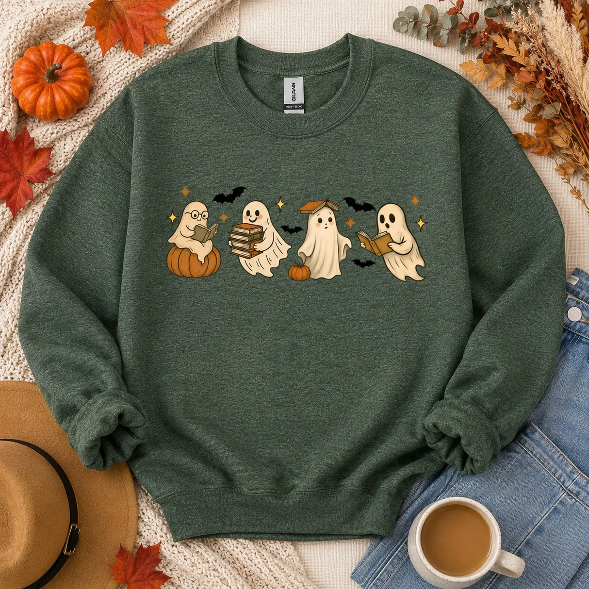 A dark heather green crewneck sweatshirt featuring a graphic of four cute cartoon ghosts in Halloween costumes, styled in a flat lay with autumn props including a hat, jeans, a coffee cup, and dried leaves.