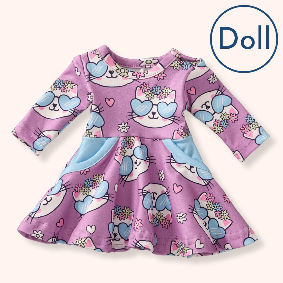 A flat lay of Pete + Lucy’s Purrfectly Cute Doll Dress, designed with long sleeves and a flared skirt. The dress is patterned with cheerful white kitten faces wearing blue heart-shaped sunglasses and floral crowns, with pink hearts and blossoms scattered across a dusty mauve background. Light blue side pockets provide a soft pop of color.