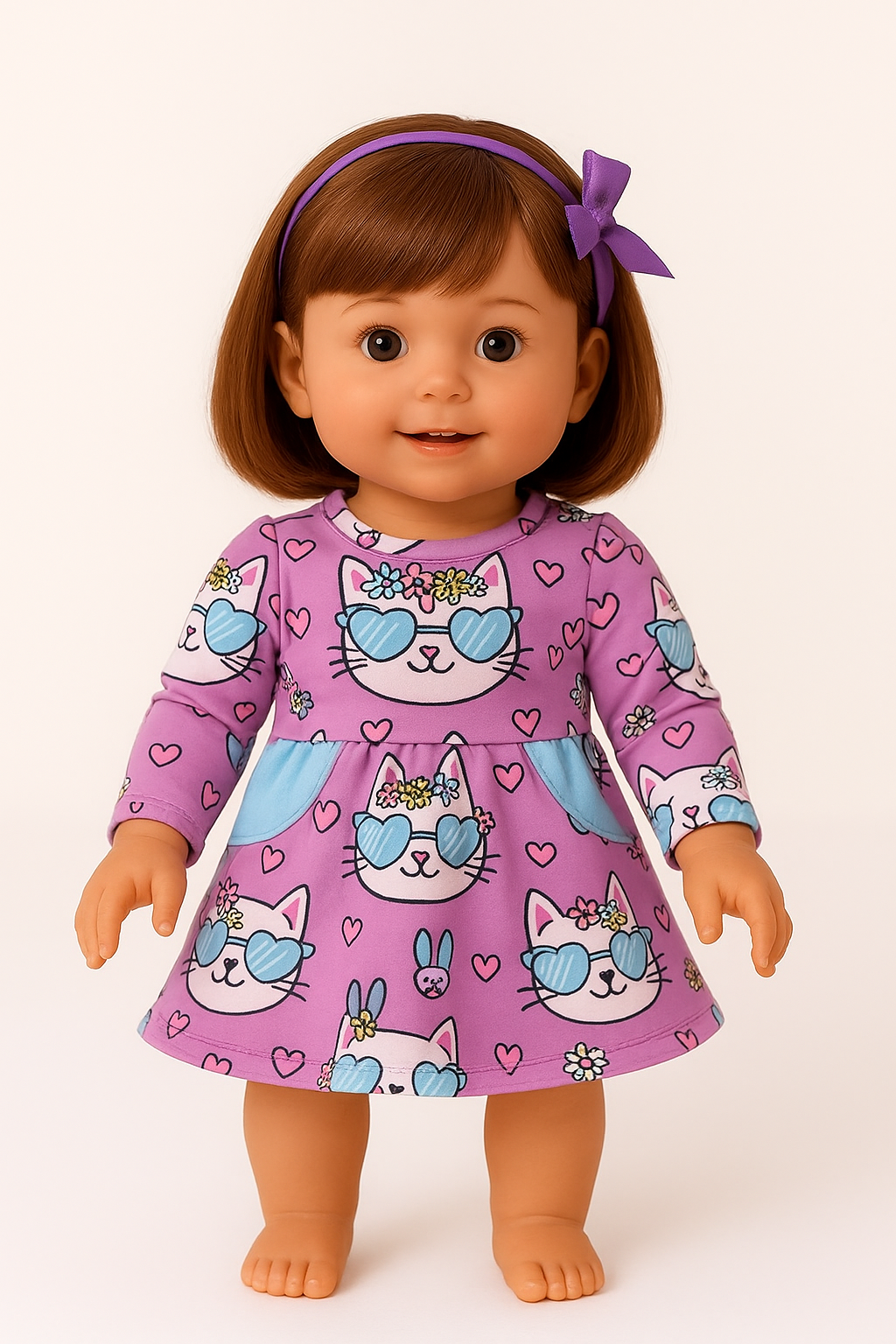 A smiling American Girl-style doll wearing Pete + Lucy’s Purrfectly Cute Doll Dress, featuring long sleeves and a flared skirt. The dress is printed with white kitten faces in blue heart-shaped sunglasses and floral crowns, scattered among pink hearts on a dusty mauve background. Light blue side pockets add a playful pop of color. The doll wears a purple bow headband and has a cheerful expression.