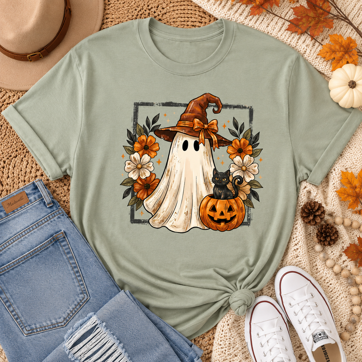 A sage green graphic t-shirt featuring a cute Halloween illustration of a ghost in a witch hat, a black cat, and pumpkins surrounded by flowers. The t-shirt is styled in a flat-lay arrangement with blue jeans, white sneakers, a brown hat, and autumn-themed accessories on a beige woven mat.