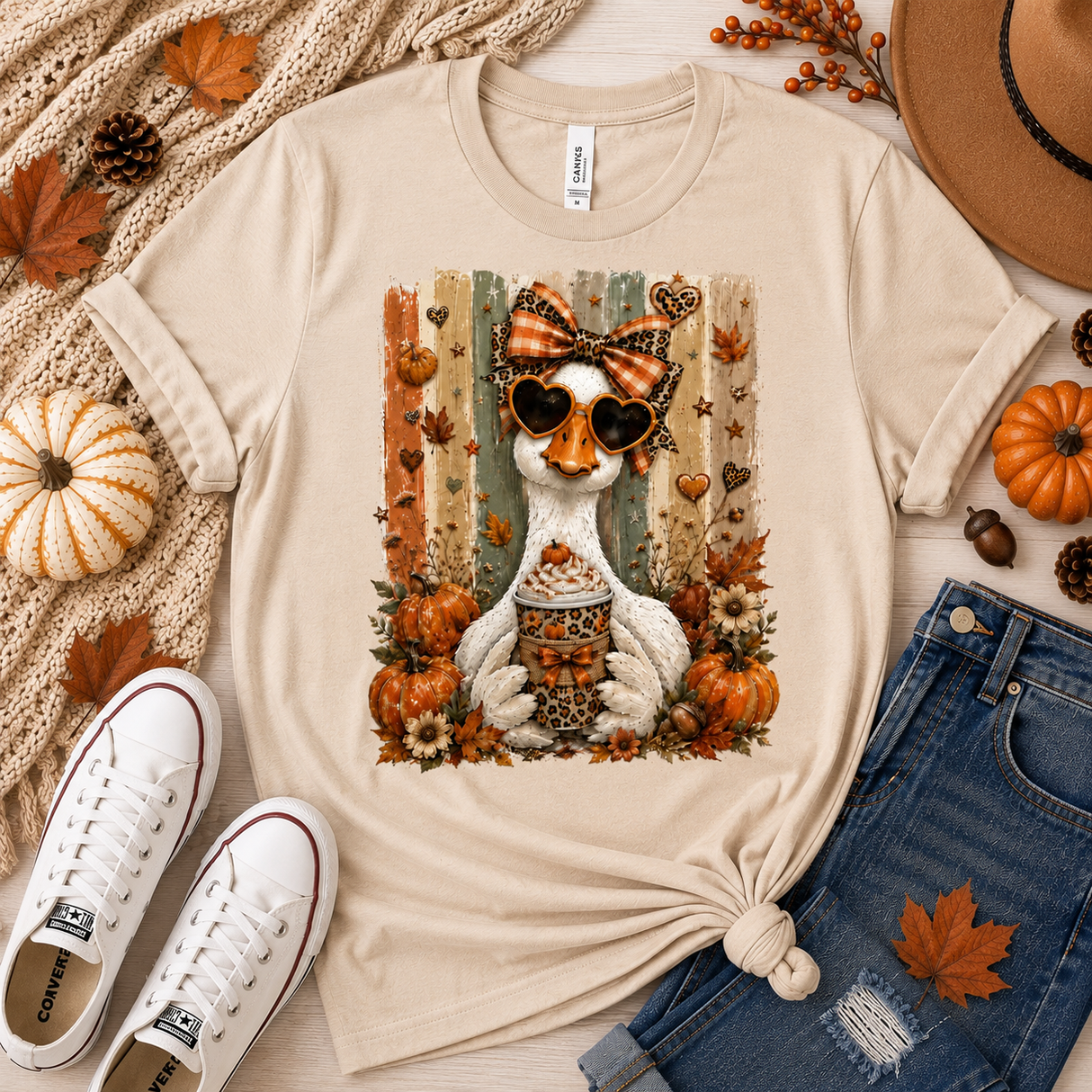 A natural color graphic t-shirt featuring a white goose wearing sunglasses and a bow, surrounded by pumpkins, autumn leaves, and a hotdog, styled in a flat lay with jeans, sneakers, a hat, and fall decor.