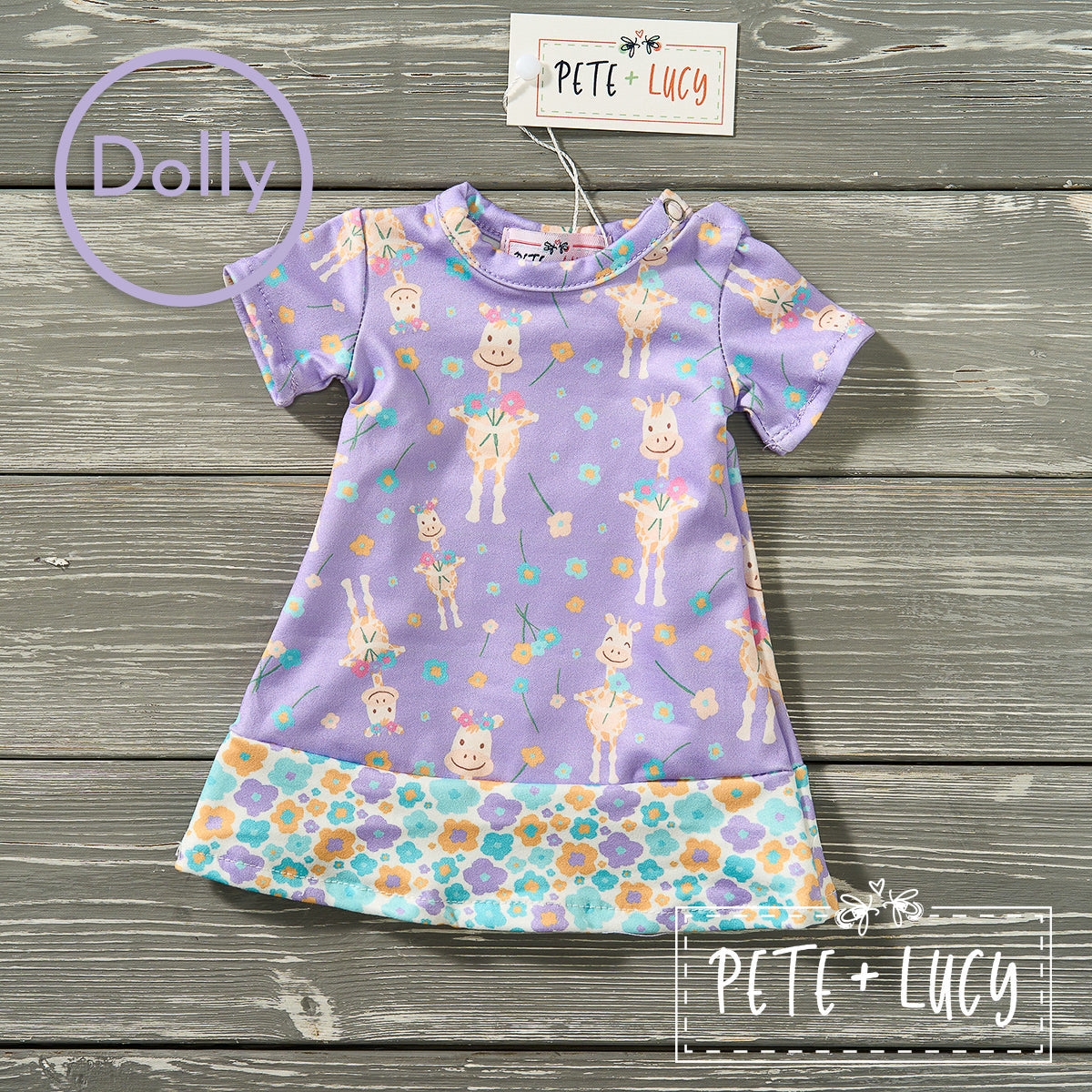 Flat lay of a purple Pete + Lucy dolly dress with cheerful giraffes and floral trim, displayed on a wood background.