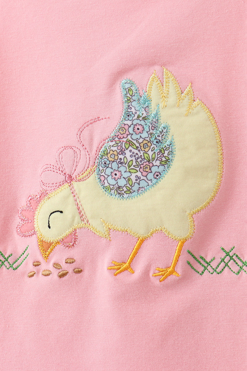 Close-up of a yellow chick appliqué with floral-patterned wings, embroidered bow, orange feet, and scattered seeds on pink fabric.