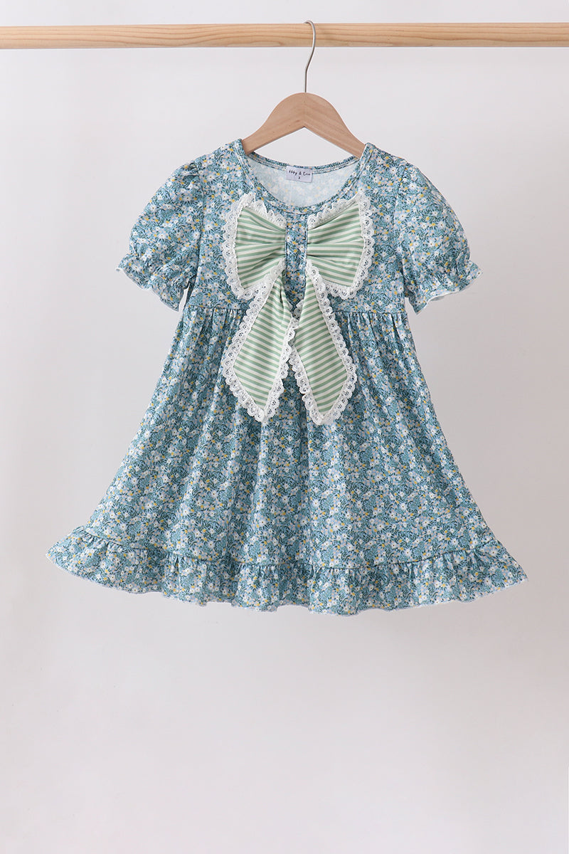 Girls’ blue floral dress with puff sleeves, a ruffled hem, and an oversized sage-striped bow trimmed in white lace.