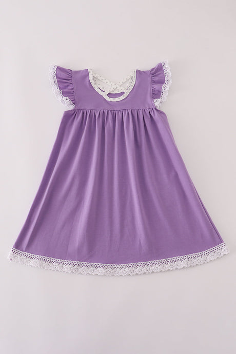 A flat lay of the back side of a girls' purple dress made from soft milk silk fabric. The dress features flutter sleeves trimmed in white lace, a scooped neckline with polka-dot lining, and a charming crisscross strap detail at the upper back. The hem is finished with delicate white lace, adding a sweet touch to this comfortable, twirl-ready Halloween look.