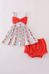 A flat lay of a baby girl outfit set featuring a sleeveless sundress with wide shoulder straps. The dress has a blue-and-white striped background with vertical rows of red apples and greenery, and a large red bow centered on the chest. It is paired with solid red bloomers that have gathered elastic at the legs for a comfortable fit.