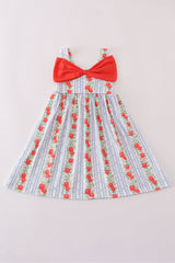 A flat lay of a girls’ sleeveless sundress featuring wide shoulder straps and a flared skirt. The dress has a blue-and-white striped background with vertical rows of red apples and leafy vines. A large red bow decorates the front chest area, adding a playful touch to the classic print. The design is sweet and vintage-inspired, perfect for warm weather.