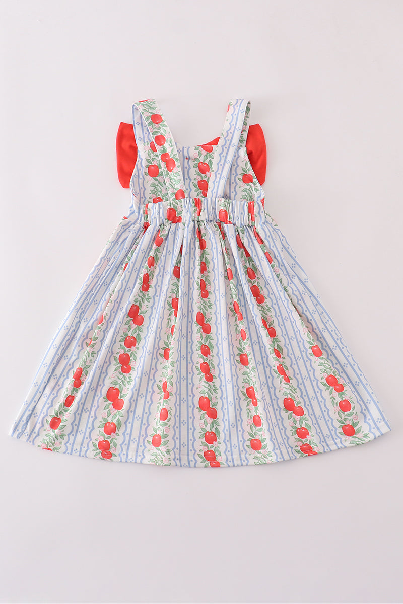 Back view of a girls’ sleeveless sundress featuring wide straps and an elastic waistband. The dress showcases a vintage-style print with vertical rows of red apples and leafy vines on a soft blue-and-white striped background. The red bow from the front peeks out slightly at the sides. The gathered waist and full skirt create a classic, twirl-ready silhouette.
