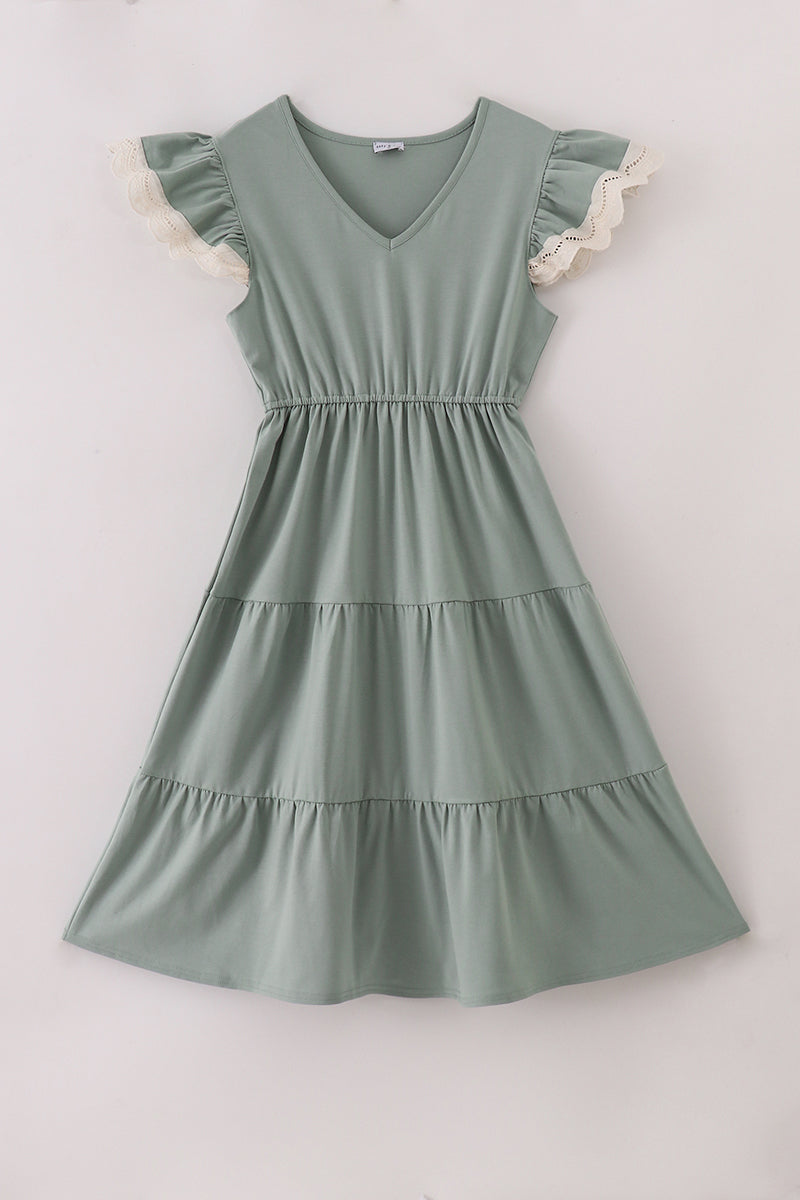 A sage green tiered dress with a V-neckline and ruffled flutter sleeves trimmed in delicate ivory lace, laid flat on a light background. The dress features an elastic waistband and three gathered tiers for a soft, flowy silhouette.