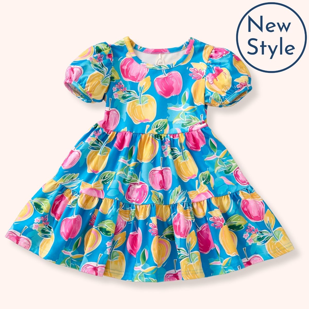 A flat lay of the Pete + Lucy A+ Apples Dress, styled with short puff sleeves and a tiered silhouette. The vibrant turquoise background is decorated with whimsical yellow and pink apples, leafy details, and sweet bows.