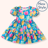 A flat lay of the Pete + Lucy A+ Apples Dress, styled with short puff sleeves and a tiered silhouette. The vibrant turquoise background is decorated with whimsical yellow and pink apples, leafy details, and sweet bows.