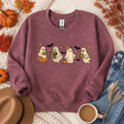 A mauve crewneck sweatshirt with a graphic of four cute cartoon ghosts reading books, styled in a flat lay with autumn leaves, pumpkins, a hat, jeans, and a cup of coffee.