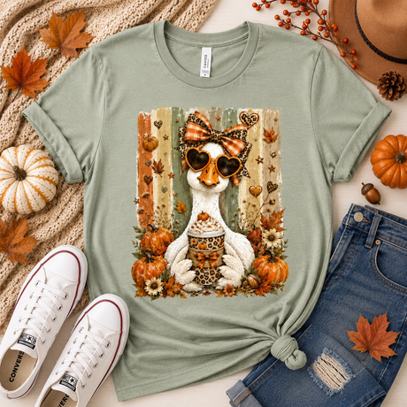 A sage green graphic t-shirt featuring a white chicken wearing heart-shaped sunglasses and a leopard-print bow, surrounded by autumn elements like pumpkins, leaves, and a hotdog with a drink. The shirt is styled in a flat lay with white Converse sneakers, blue jeans, a knit sweater, and fall leaves.