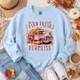 A light blue crewneck sweatshirt with a colorful graphic of a pink pickup truck loaded with pumpkins and the words 'FARM FRESH PUMPKINS'. The sweatshirt is styled in a flat lay with a hat, jeans, white sneakers, a coffee cup, and autumn leaves.
