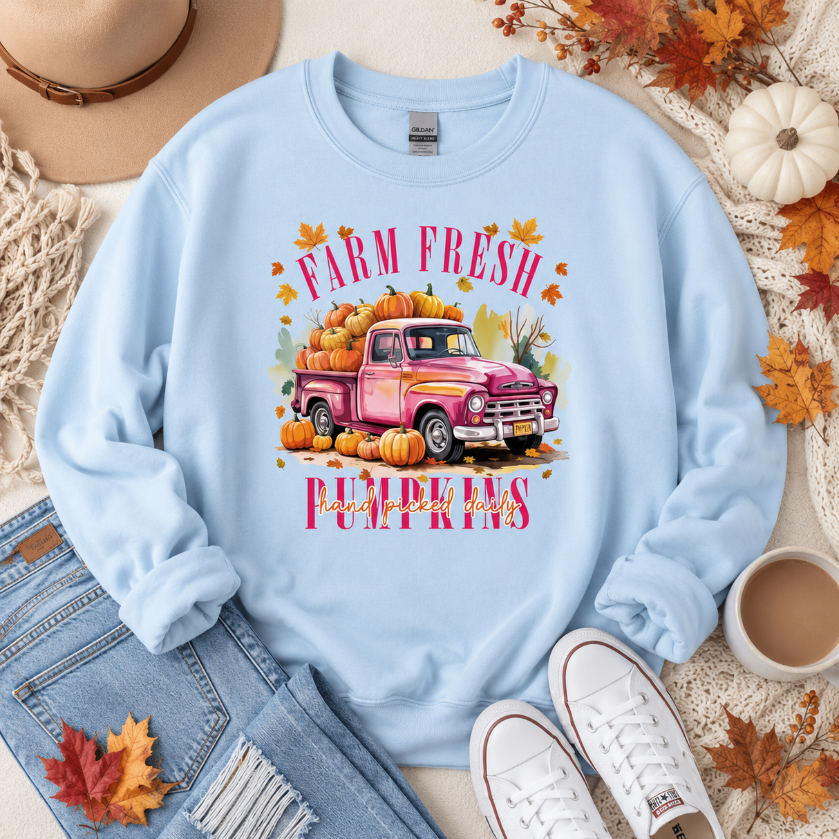 A light blue crewneck sweatshirt with a colorful graphic of a pink pickup truck loaded with pumpkins and the words 'FARM FRESH PUMPKINS'. The sweatshirt is styled in a flat lay with a hat, jeans, white sneakers, a coffee cup, and autumn leaves.