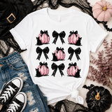 A white graphic t-shirt featuring a 3x3 grid of pink pumpkins, black bows, and black cats, styled in a flat-lay with jeans, black lace, black sneakers, and decorative pumpkins.