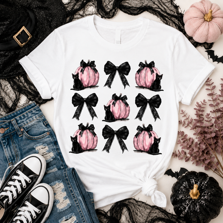A white graphic t-shirt featuring a 3x3 grid of pink pumpkins, black bows, and black cats, styled in a flat-lay with jeans, black lace, black sneakers, and decorative pumpkins.