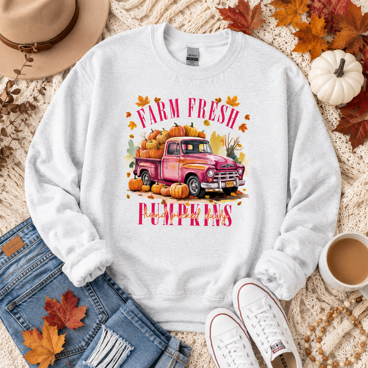 A white crewneck sweatshirt laid flat on a cozy autumn-themed background. The sweatshirt features a colorful graphic of a pink vintage pickup truck loaded with pumpkins, with the text 'FARM FRESH' above and 'PUMPKINS' below in pink.