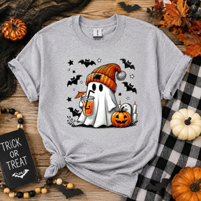 A heather gray short-sleeve t-shirt with a Halloween graphic of a ghost in an orange beanie holding a pumpkin drink, surrounded by bats, a jack-o'-lantern, and candles. The shirt is styled in a flat lay on a wooden surface with autumn props including pumpkins, a buffalo plaid blanket, and a 'Trick or Treat' sign.