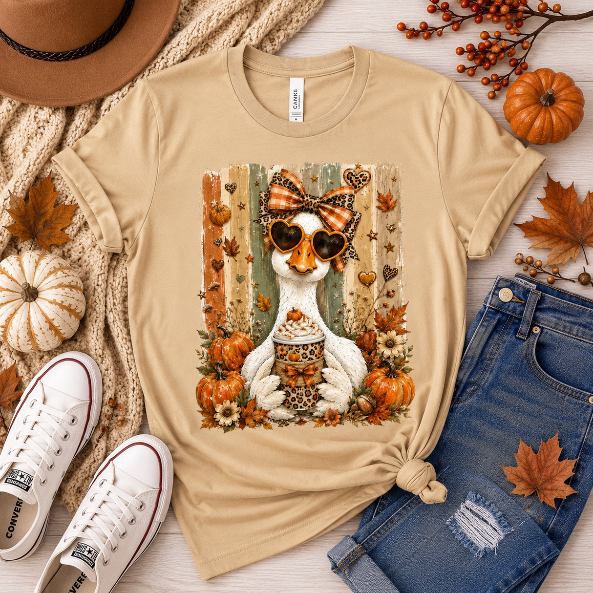 A beige graphic t-shirt featuring a white duck wearing sunglasses and a bow tie with autumn elements, styled in a flat lay with jeans, sneakers, a hat, and fall leaves on a light wood background.