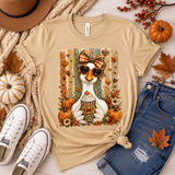 A beige graphic t-shirt featuring a white duck wearing sunglasses and a bow tie with autumn elements, styled in a flat lay with jeans, sneakers, a hat, and fall leaves on a light wood background.