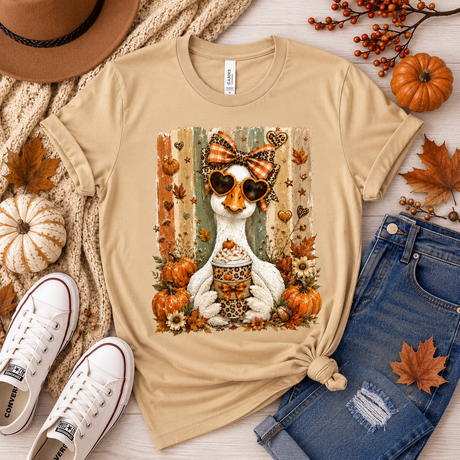 A beige graphic t-shirt featuring a white duck wearing sunglasses and a bow tie with autumn elements, styled in a flat lay with jeans, sneakers, a hat, and fall leaves on a light wood background.