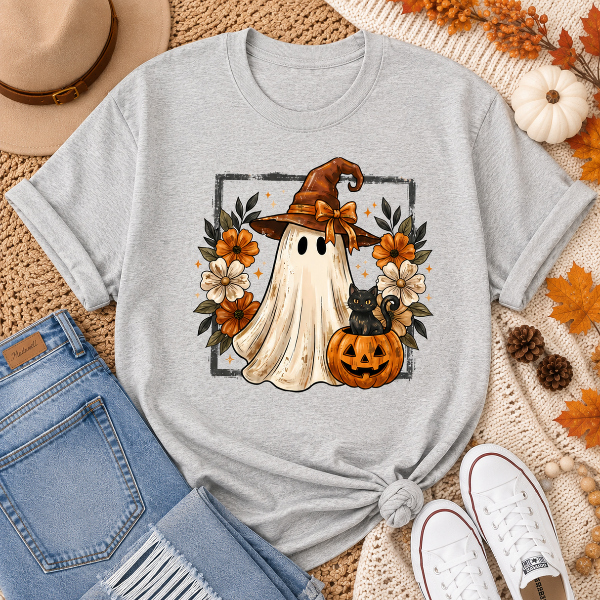 A light gray short-sleeve T-shirt laid flat with a Halloween graphic featuring a ghost, witch hat, black cat, pumpkin, and autumn flowers. The shirt is styled with blue jeans, white sneakers, a tan hat, and fall leaves on a cream woven rug.