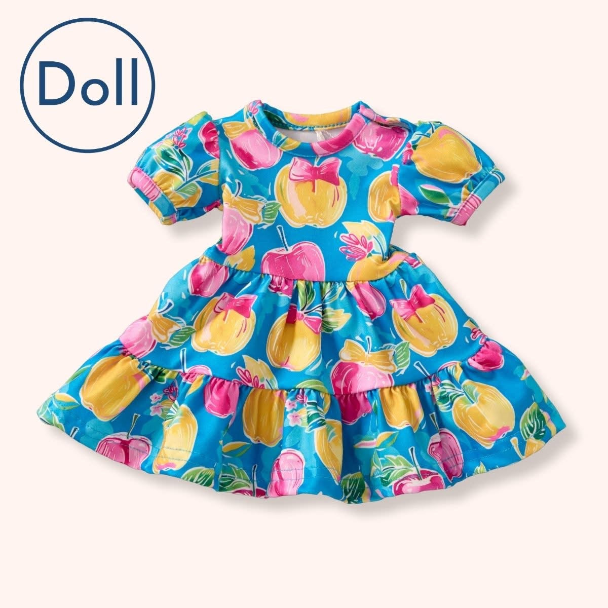 A flat lay of the Pete + Lucy A+ Apples Doll Dress, perfectly matching the child-size version with its puff sleeves and tiered design. Features bright yellow and pink apples with bows on a turquoise blue background.