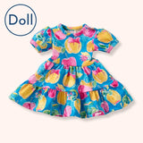 A flat lay of the Pete + Lucy A+ Apples Doll Dress, perfectly matching the child-size version with its puff sleeves and tiered design. Features bright yellow and pink apples with bows on a turquoise blue background.