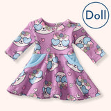 A flat lay of Pete + Lucy’s Purrfectly Cute Doll Dress, designed with long sleeves and a flared skirt. The dress is patterned with cheerful white kitten faces wearing blue heart-shaped sunglasses and floral crowns, with pink hearts and blossoms scattered across a dusty mauve background. Light blue side pockets provide a soft pop of color.
