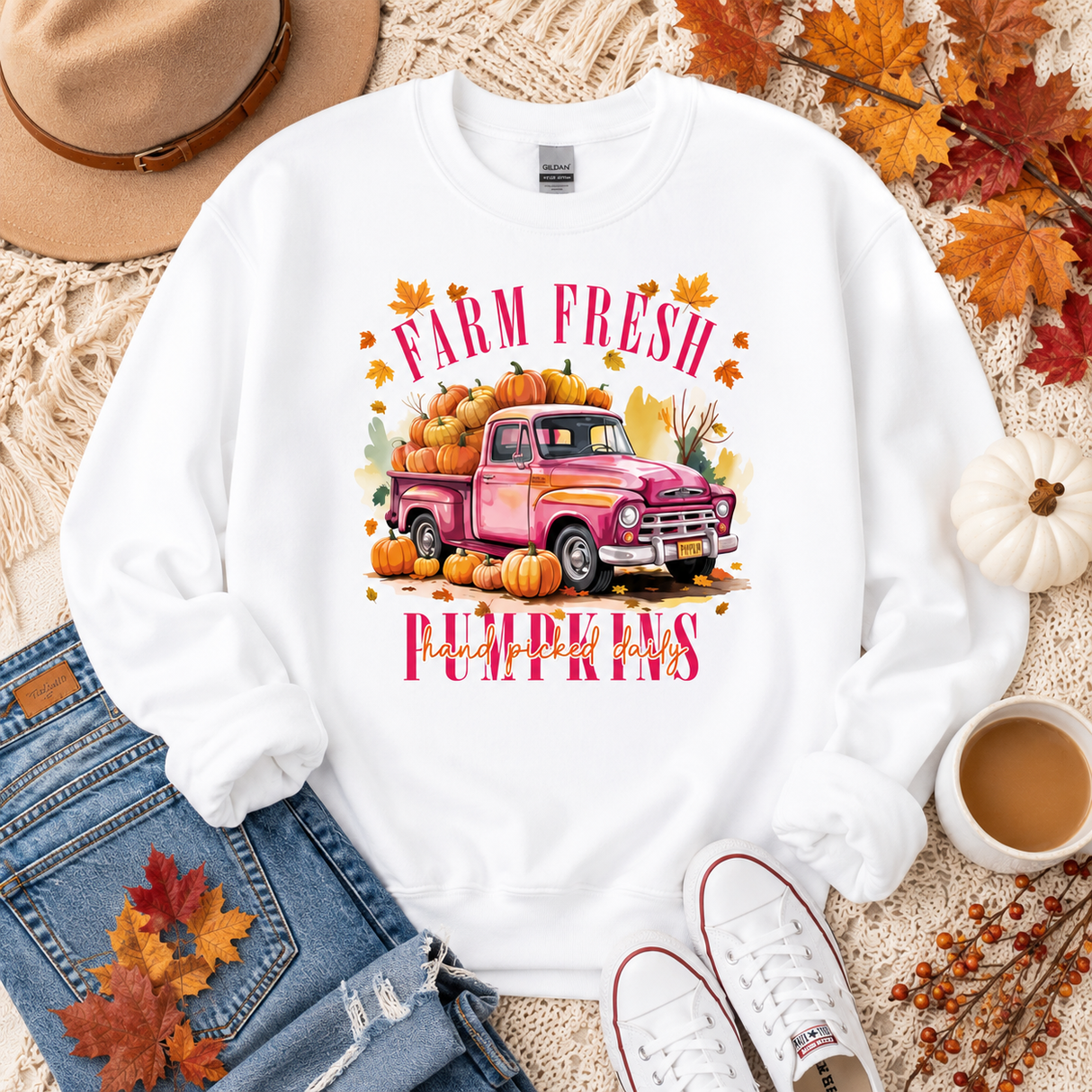 A white crewneck sweatshirt with a colorful graphic of a pink pickup truck loaded with pumpkins and the words 'FARM FRESH PUMPKINS' in pink lettering. The sweatshirt is styled in a flat lay with blue jeans, a tan hat, white sneakers, a cup of coffee, and autumn leaves on a beige knit blanket.