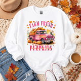 A white crewneck sweatshirt with a colorful graphic of a pink pickup truck loaded with pumpkins and the words 'FARM FRESH PUMPKINS' in pink lettering. The sweatshirt is styled in a flat lay with blue jeans, a tan hat, white sneakers, a cup of coffee, and autumn leaves on a beige knit blanket.