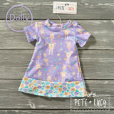 Flat lay of a purple Pete + Lucy dolly dress with cheerful giraffes and floral trim, displayed on a wood background.