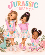 Three storybook illustrated young girls wearing matching pink and floral 'Jurassic Dreams' outfits pose together in a playful studio scene with dinosaur toys and pastel decorations. The text 'JURASSIC DREAMS' appears at the top in colorful lettering.