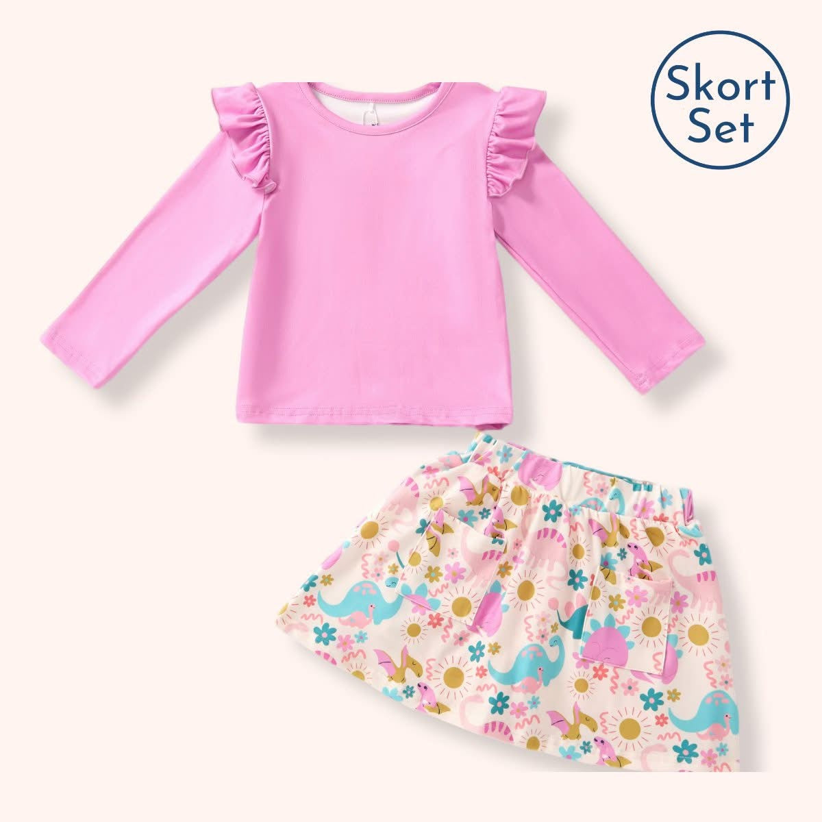 A children's two-piece outfit set featuring a pink long-sleeve top with ruffled shoulders and a matching printed skirt with colorful dinosaurs and flowers, shown on a light cream background with a 'Skort Set' label in the top right.