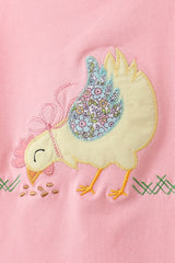 Close-up of a yellow chick appliqué with floral-patterned wings, embroidered bow, orange feet, and scattered seeds on pink fabric.