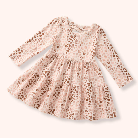 A long-sleeve baby dress with a tiered skirt, featuring a light pink and beige leopard print pattern with brown spots, shown on a solid pale pink background.