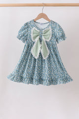 Girls’ blue floral dress with puff sleeves, a ruffled hem, and an oversized sage-striped bow trimmed in white lace.
