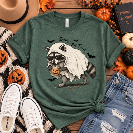Heather forest green tee featuring a raccoon in sunglasses and a ghost costume holding a jack-o’-lantern drink, with bats and “Spooky and Feral” text.