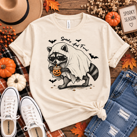 Natural cream tee featuring a raccoon in sunglasses and a ghost costume holding a jack-o’-lantern drink, with bats and “Spooky and Feral” text.