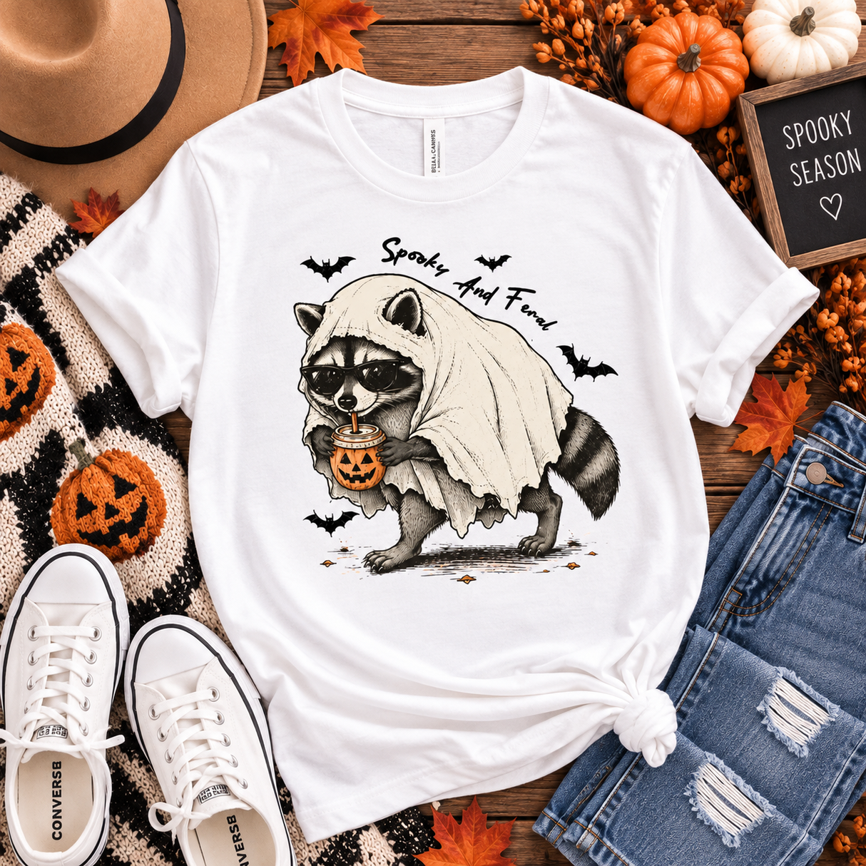 White tee featuring a raccoon in sunglasses and a ghost costume holding a jack-o’-lantern drink, with bats and “Spooky and Feral” text.