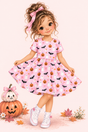 A storybook character girl with curly brown hair in a pink bow wears a pink Halloween dress with a pumpkin, bat, ghost, and bow pattern. She stands in a stylized scene with stacked pumpkins, autumn leaves, and white sneakers against a soft pink background.