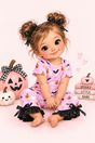 A storybook character baby with brown hair in two buns and black bows, wearing a pink Halloween pajama set with black ruffle trim, sitting on a pink background with pumpkins and books.