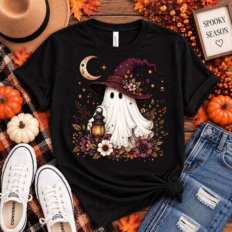 Black tee featuring a whimsical ghost in a plum witch hat holding a glowing lantern, surrounded by a crescent moon, florals, stars and fall foliage.